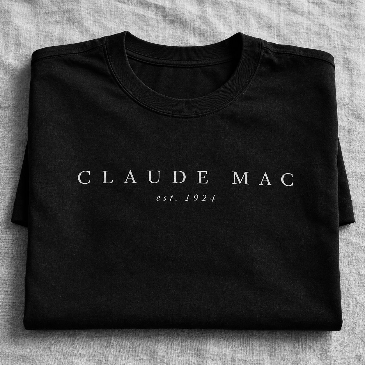 The Signature Tee, folded on linen, the Claude Mac wordmark across the chest